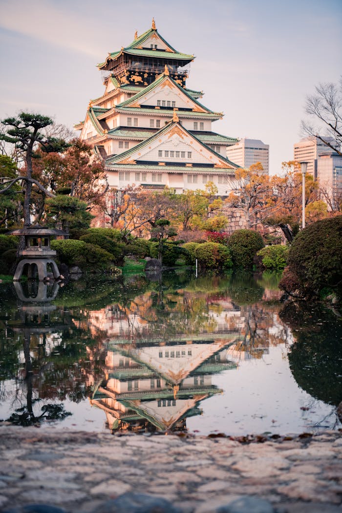 Scenic view of Osaka Castle reflected in a tranquil pond, surrounded by lush gardens at dusk.